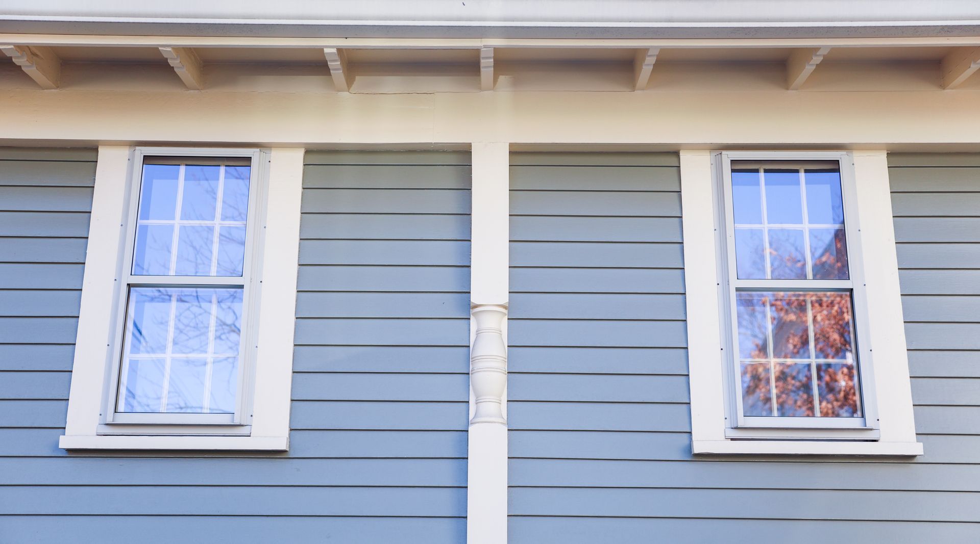 Two blue-gray windows with white trim on a blue-gray clapboard house, set against a light-colored soffit.