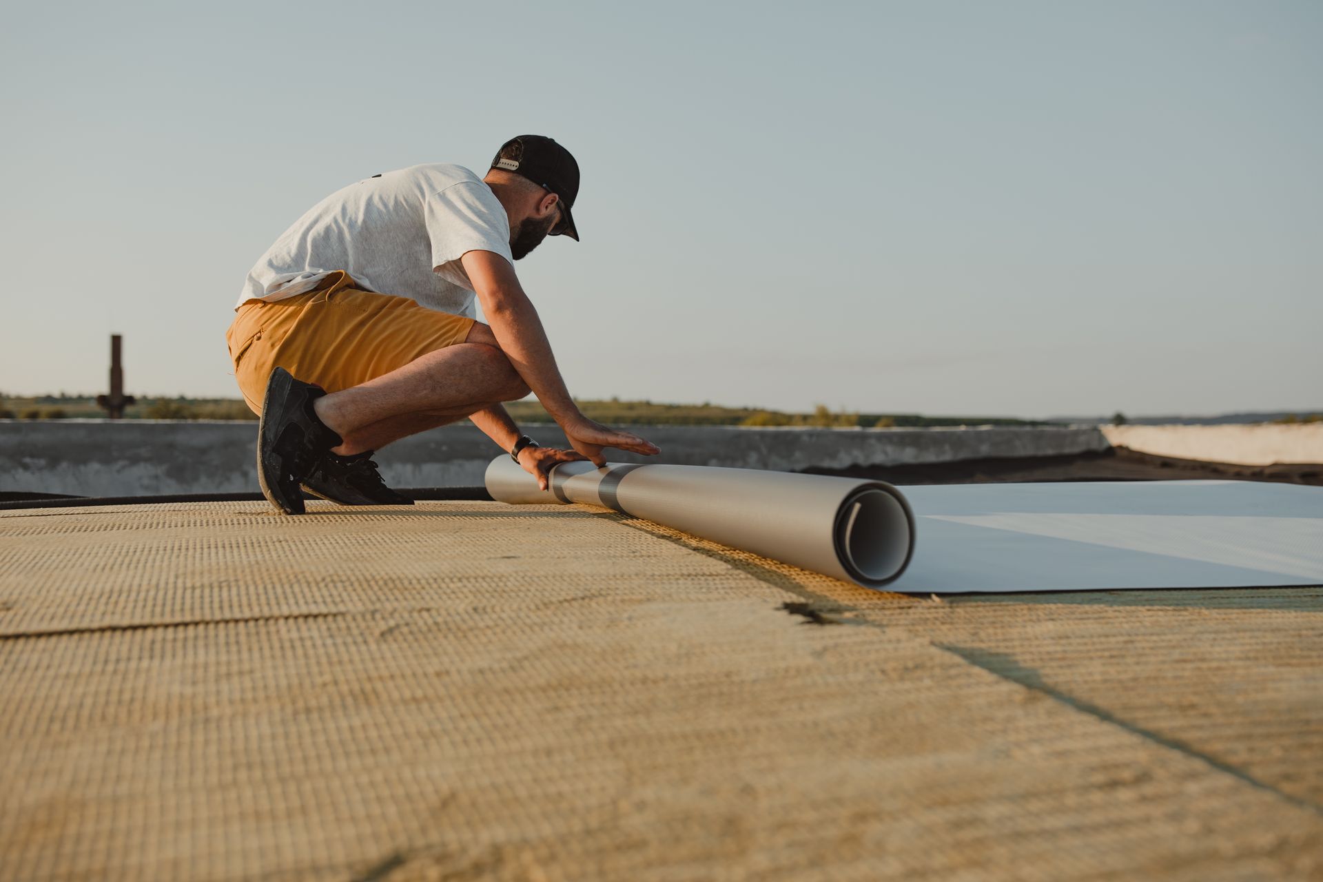 A man is kneeling down on a roof holding a roll of roofing material.