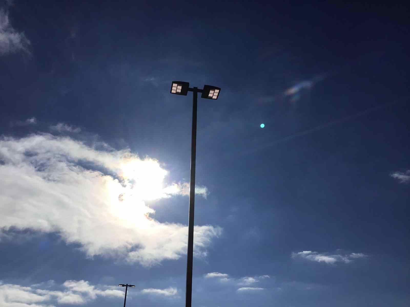 A street light against a blue sky with clouds and the sun shining through the clouds.