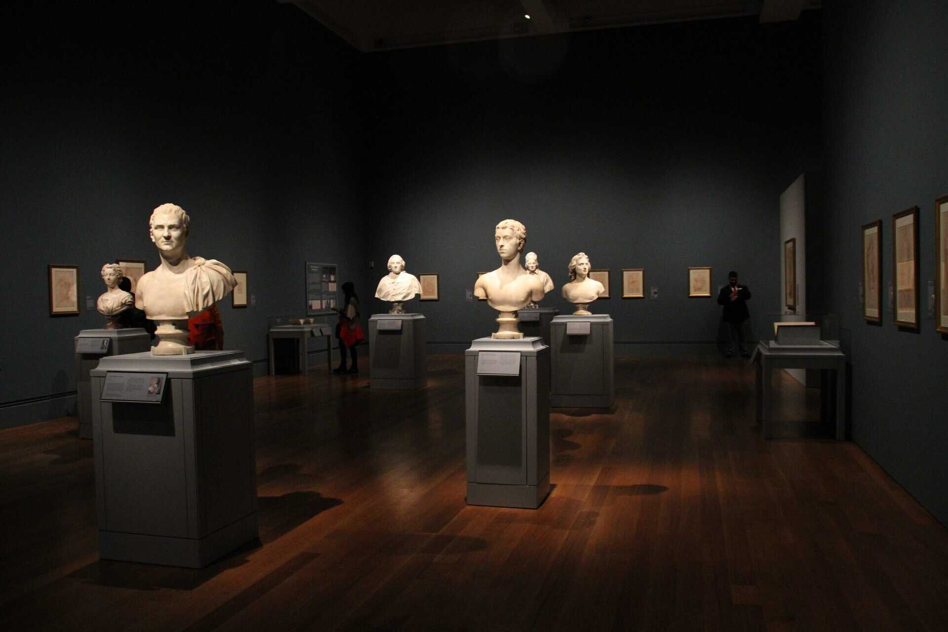Sculpture gallery with busts on pedestals and framed artwork on dark blue walls.