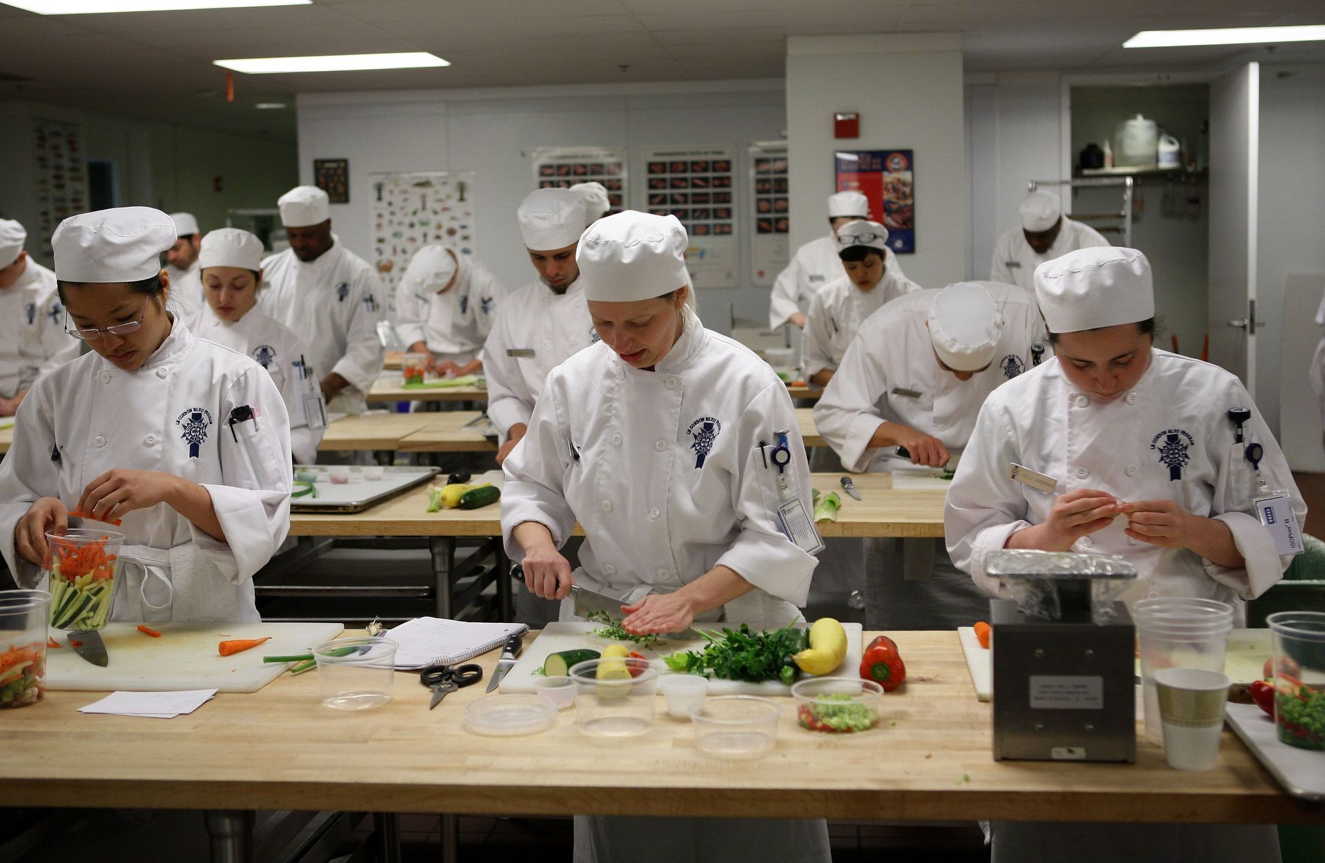 Culinary students in white chef uniforms chopping vegetables at individual workstations in a kitchen.