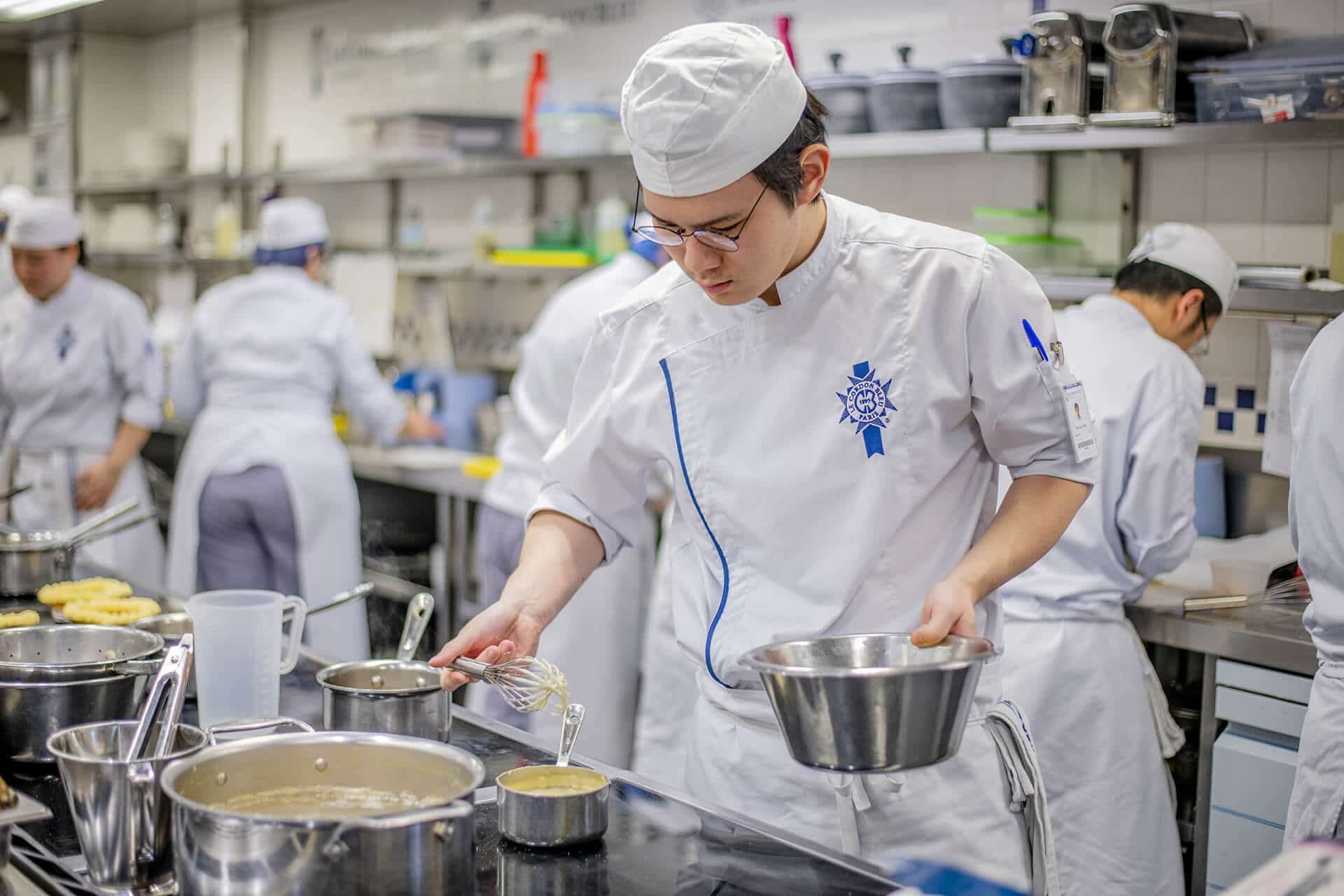Chef in a white uniform stirring food in a stainless steel bowl in a busy kitchen.