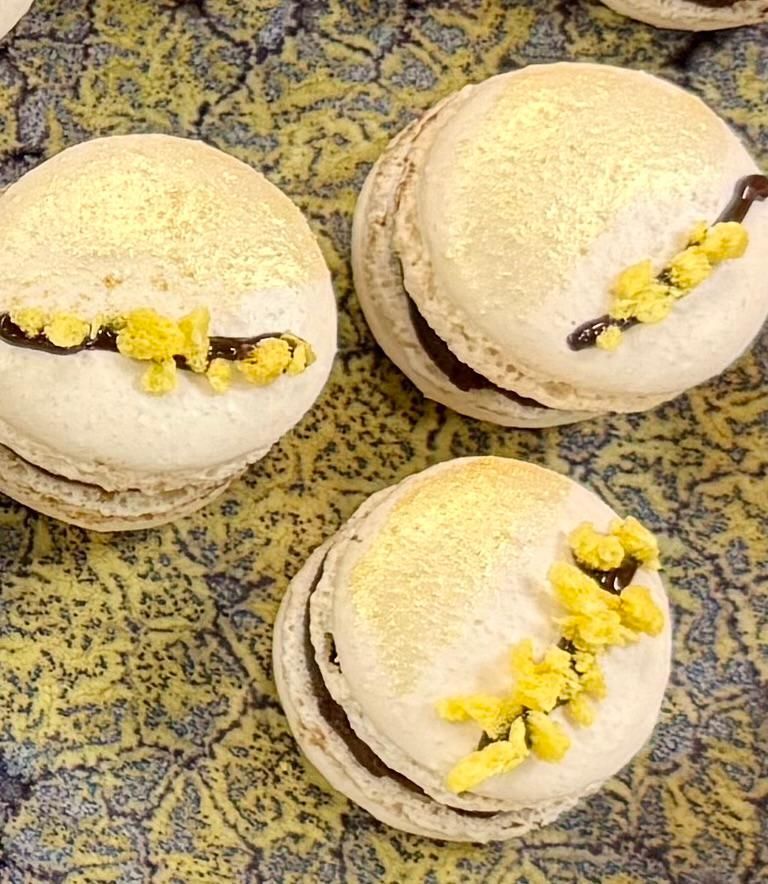 Three white macarons with chocolate filling, gold dusting, and yellow sugar decoration.