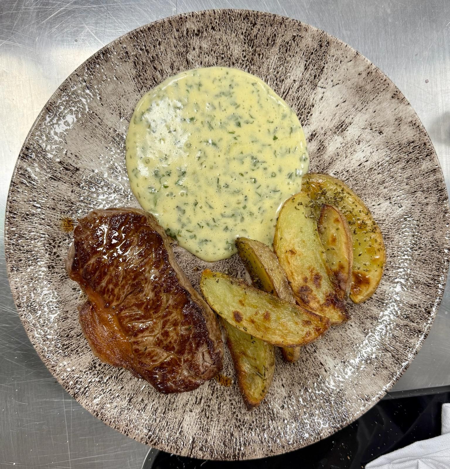 Steak with creamy sauce and potato wedges on a speckled plate.