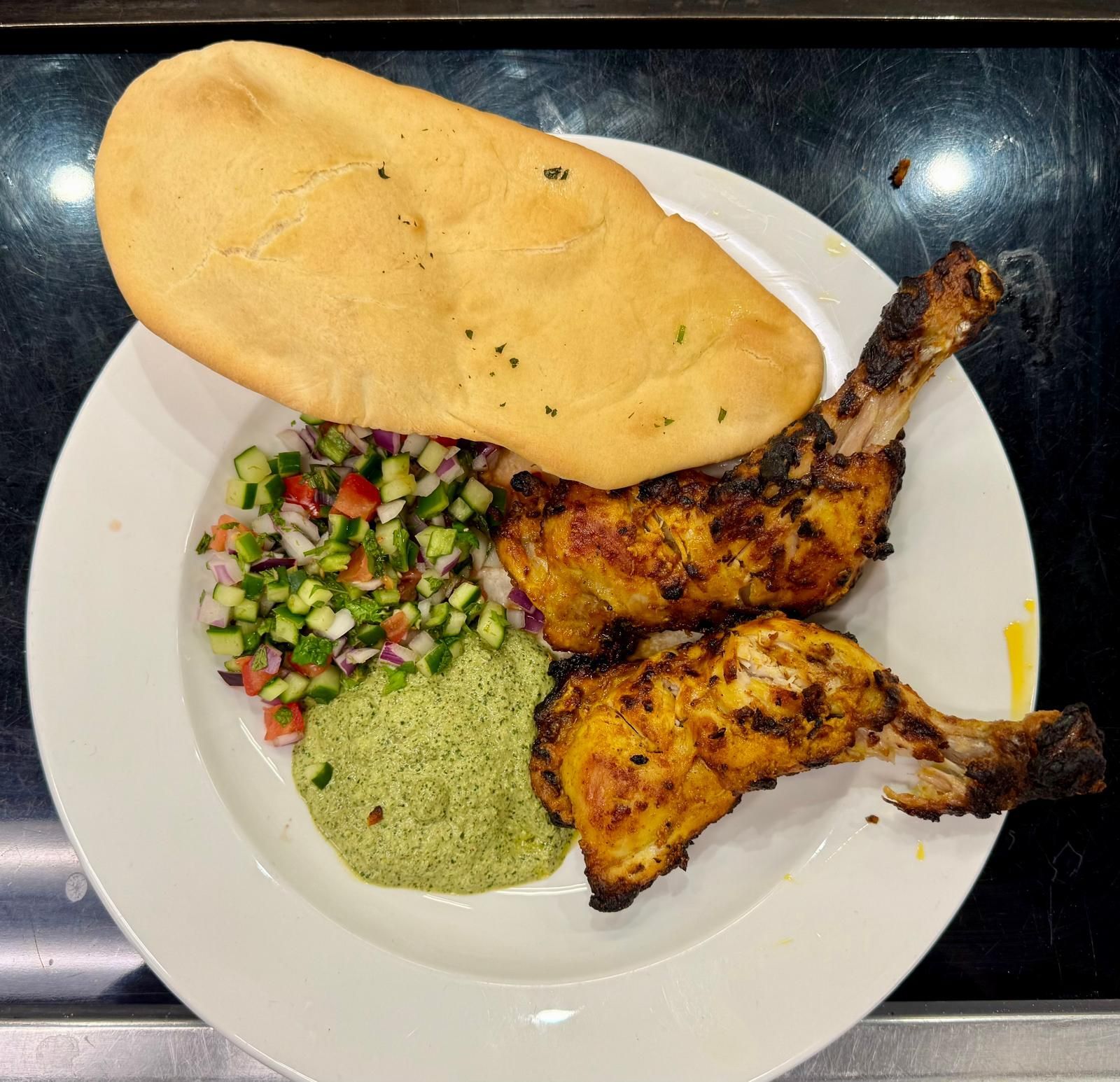 Grilled chicken legs, flatbread, cucumber salad, and green chutney on a white plate.