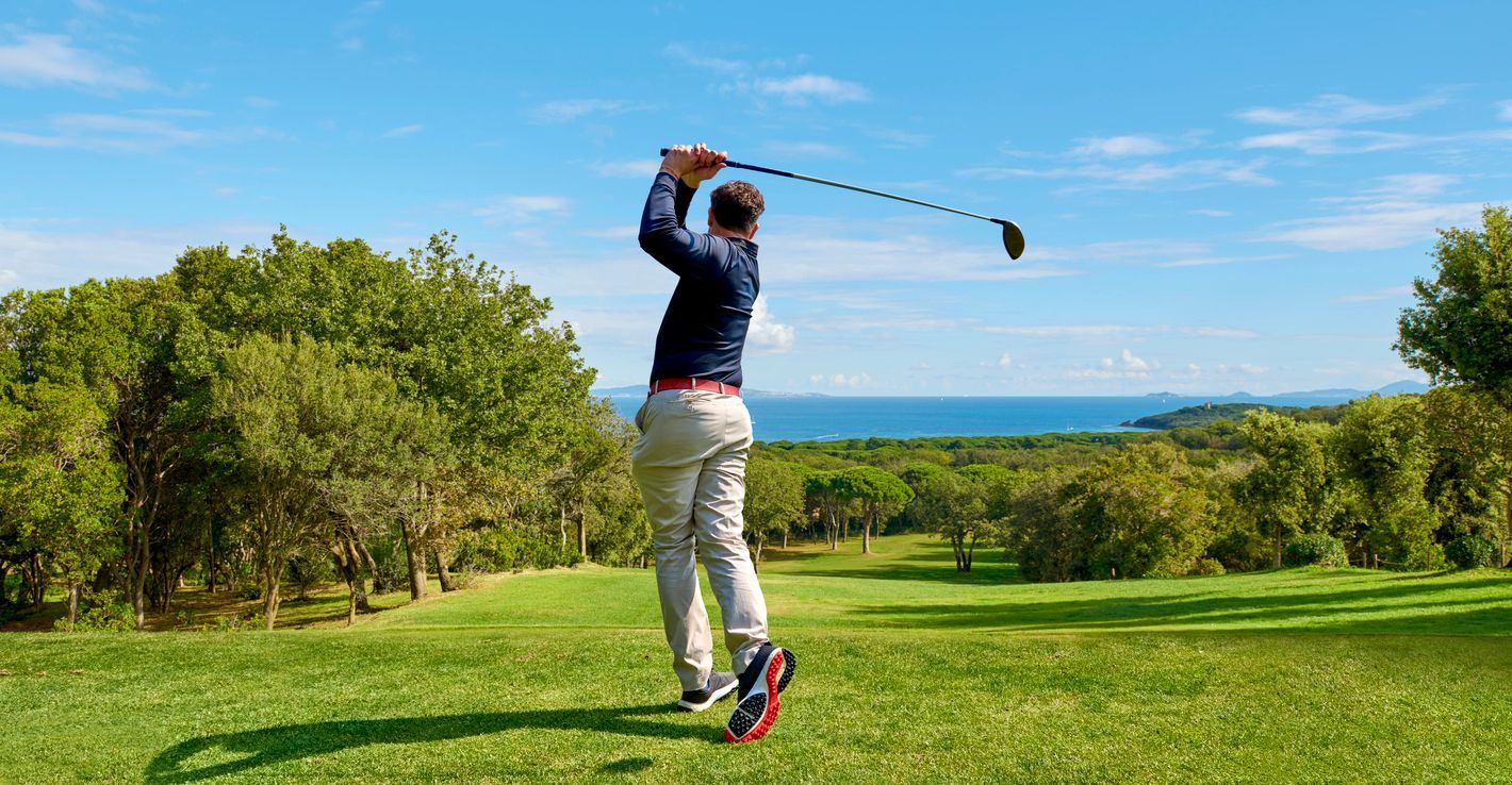 Golfer in mid-swing, swinging a golf club on a green, with trees and ocean view on a sunny day.
