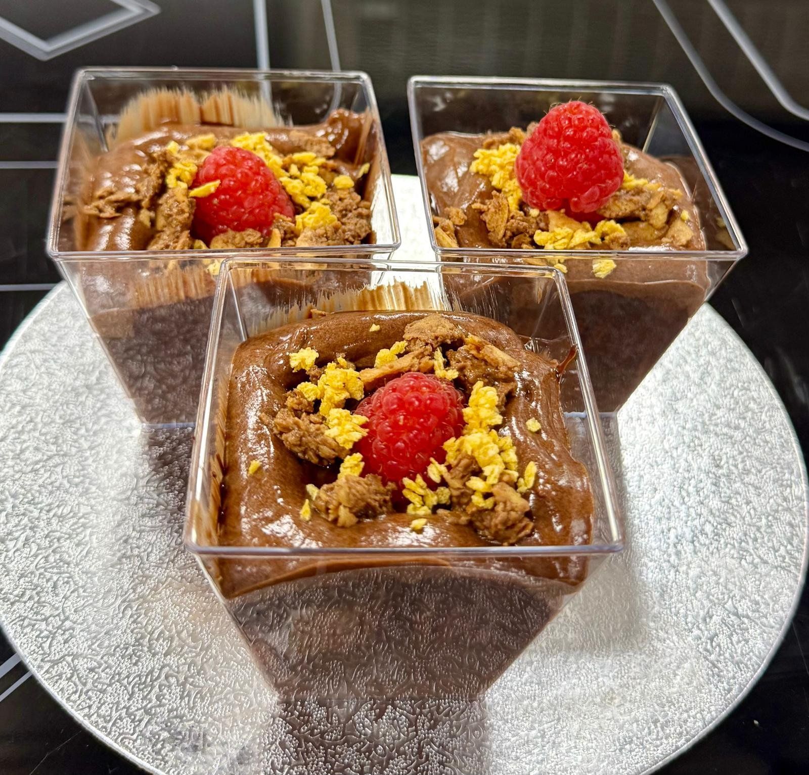 Three chocolate mousse desserts in square cups, topped with raspberries and crumbs on a silver plate.