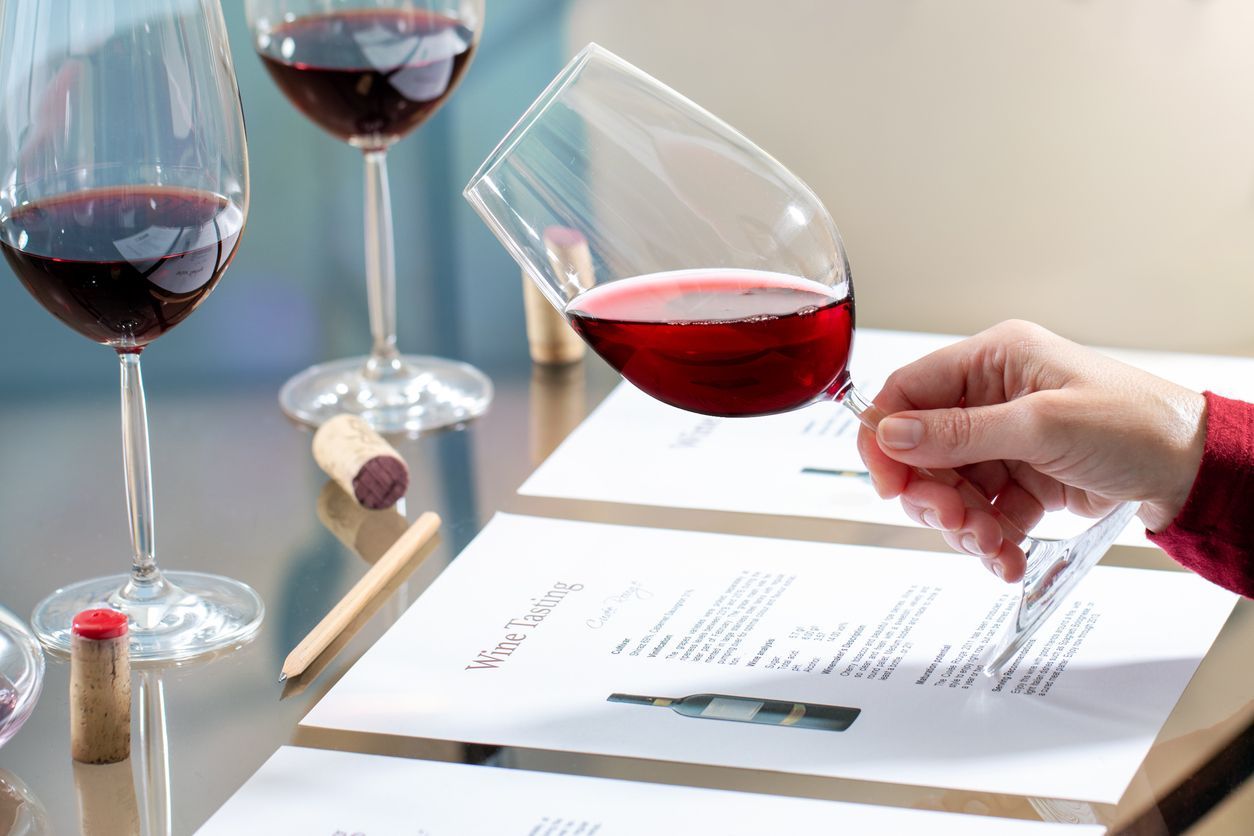A person holding a wine glass, tilting it to look at the red wine. Wine tasting setting with three glasses and papers.