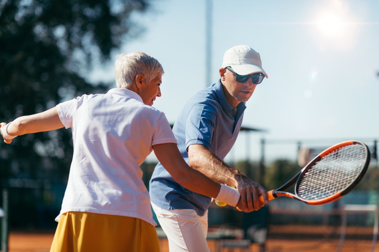 Older man receiving tennis lesson on clay court. Woman demonstrates proper swing.