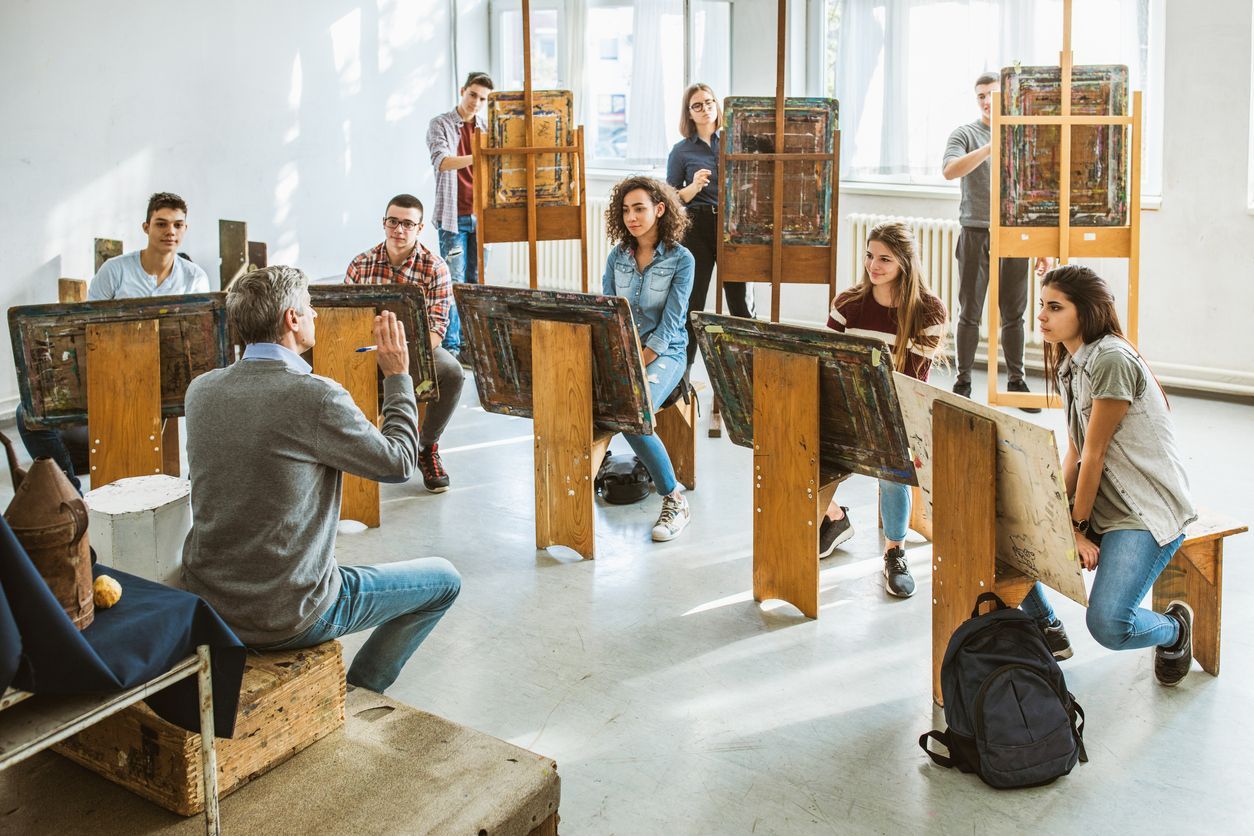 Art class: Instructor speaks to students observing their paintings on easels in a sunlit studio.