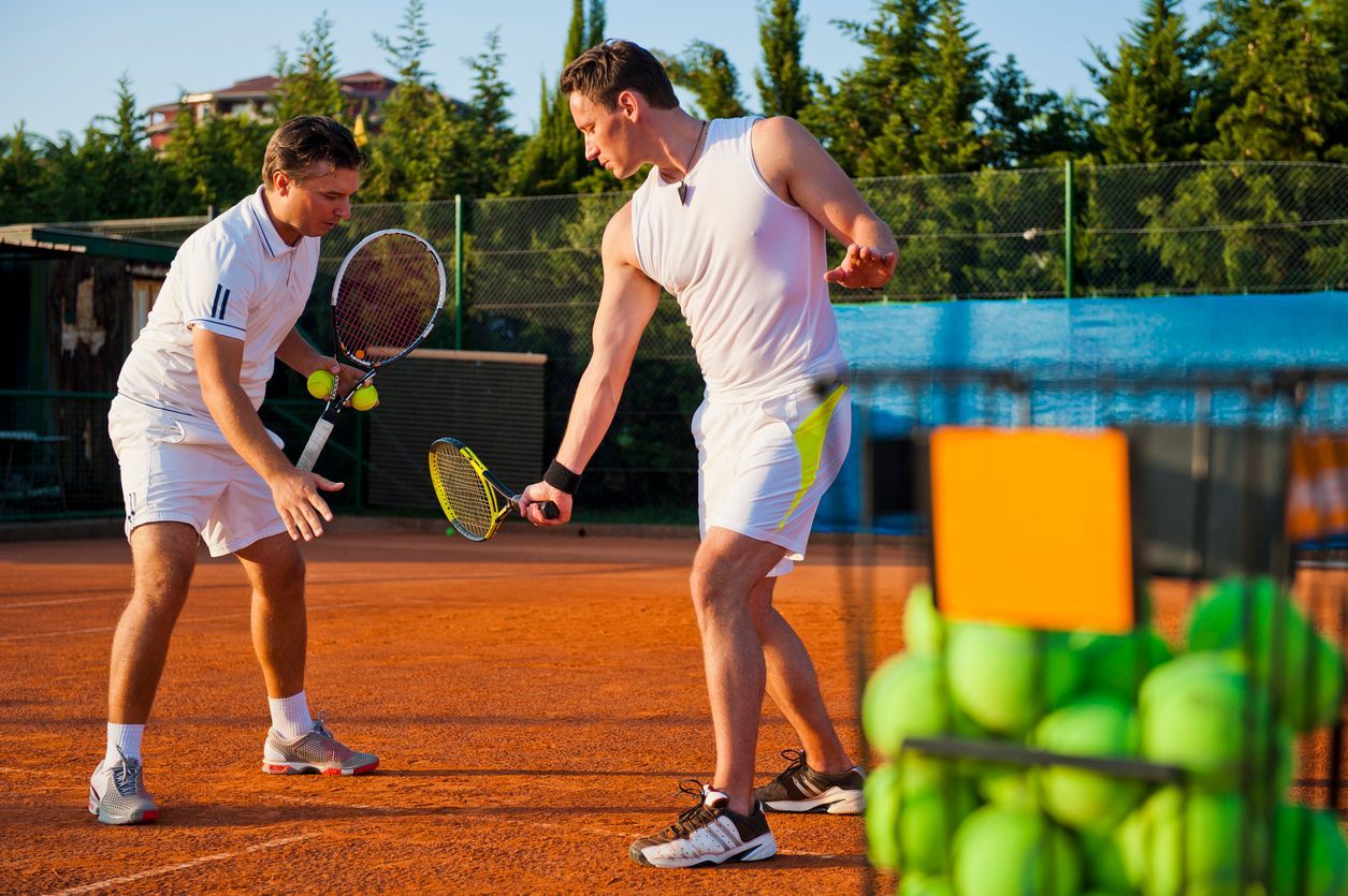 Two men on a clay tennis court: one giving a lesson, the other ready to hit. Green balls sit in a cart.