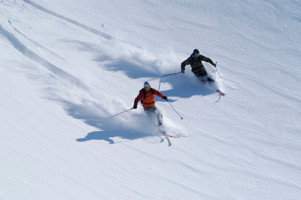 Two skiers carve down a snow-covered slope, kicking up powder. Sunny day.