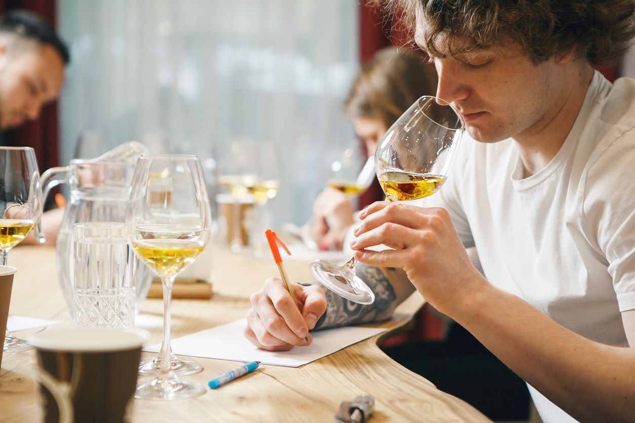 Man sniffs amber liquid in glass, writing notes. Others taste at a table.