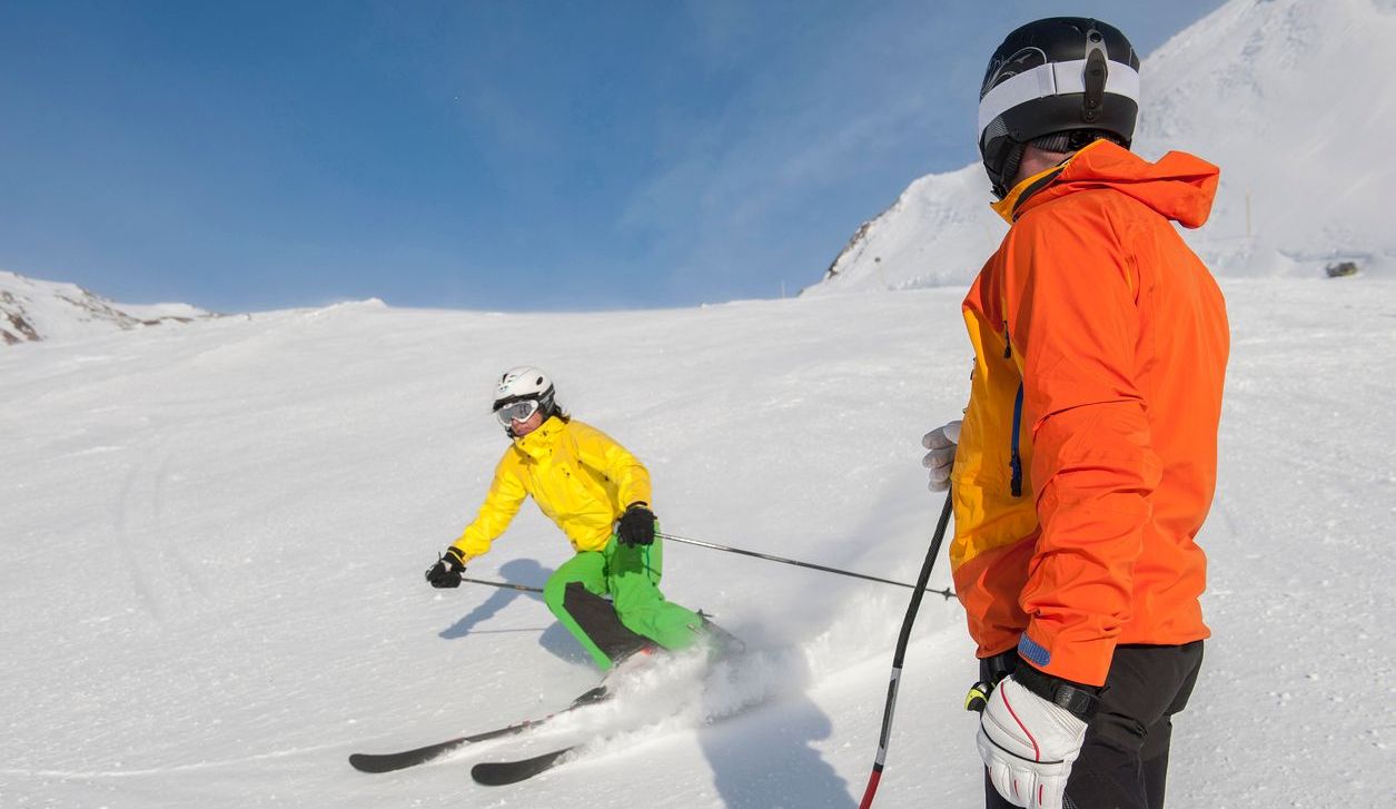 Skier in yellow jacket on snowy slope, observed by person in orange jacket.