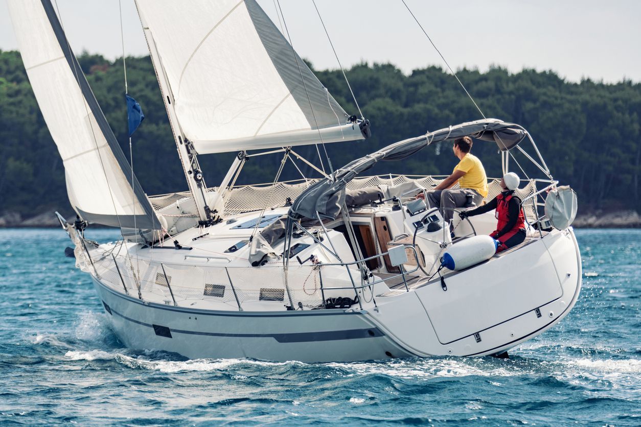 Sailboat on the water with two people on board, white sails, blue sea, green trees in the background.