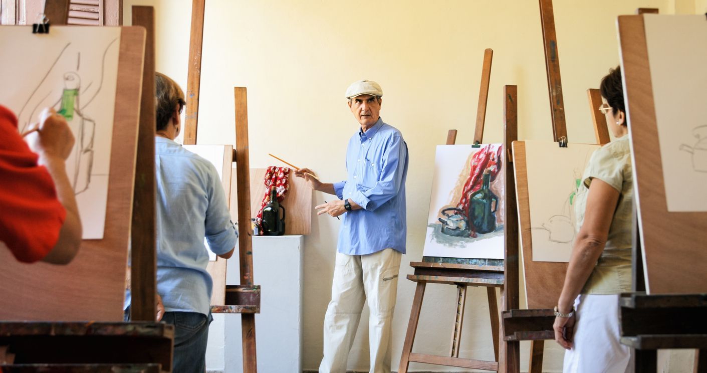 Art class: Instructor in blue shirt gestures toward a student's drawing, others at easels with their work, light-filled room.