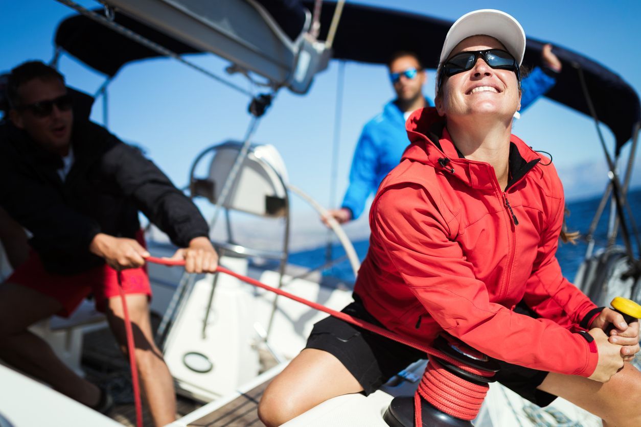 Three people sailing, pulling ropes on a boat. Woman in red jacket smiles; blue sky.