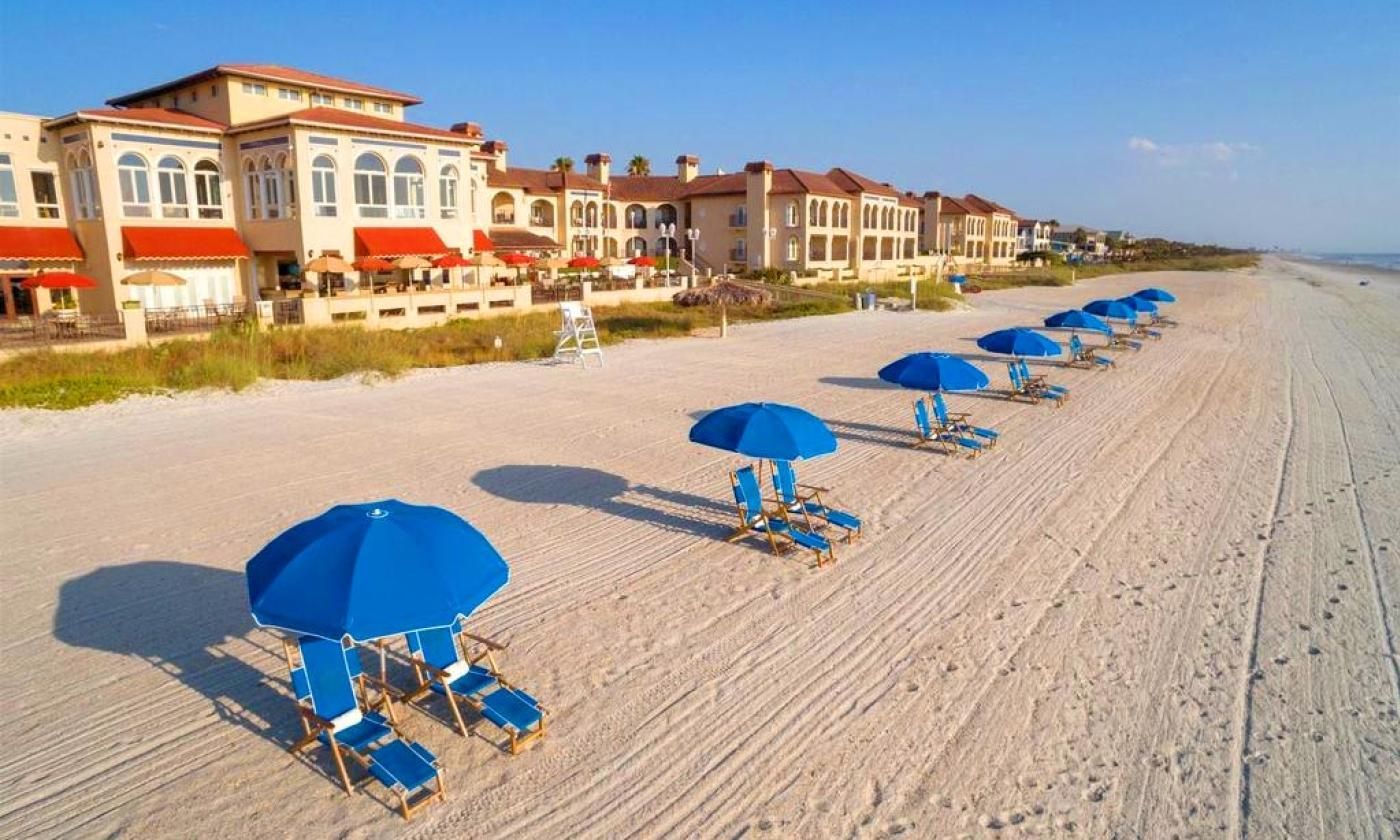 Beach with blue umbrellas and lounge chairs in front of a resort building on a sunny day.