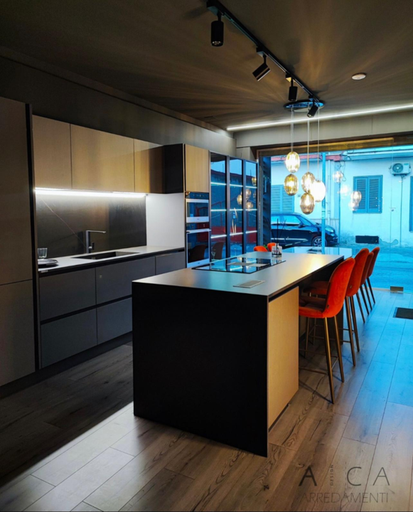 Showroom di cucine moderne con mobili grigio scuro, grande isola con posti a sedere, lampade a sospensione e sedie rosse.