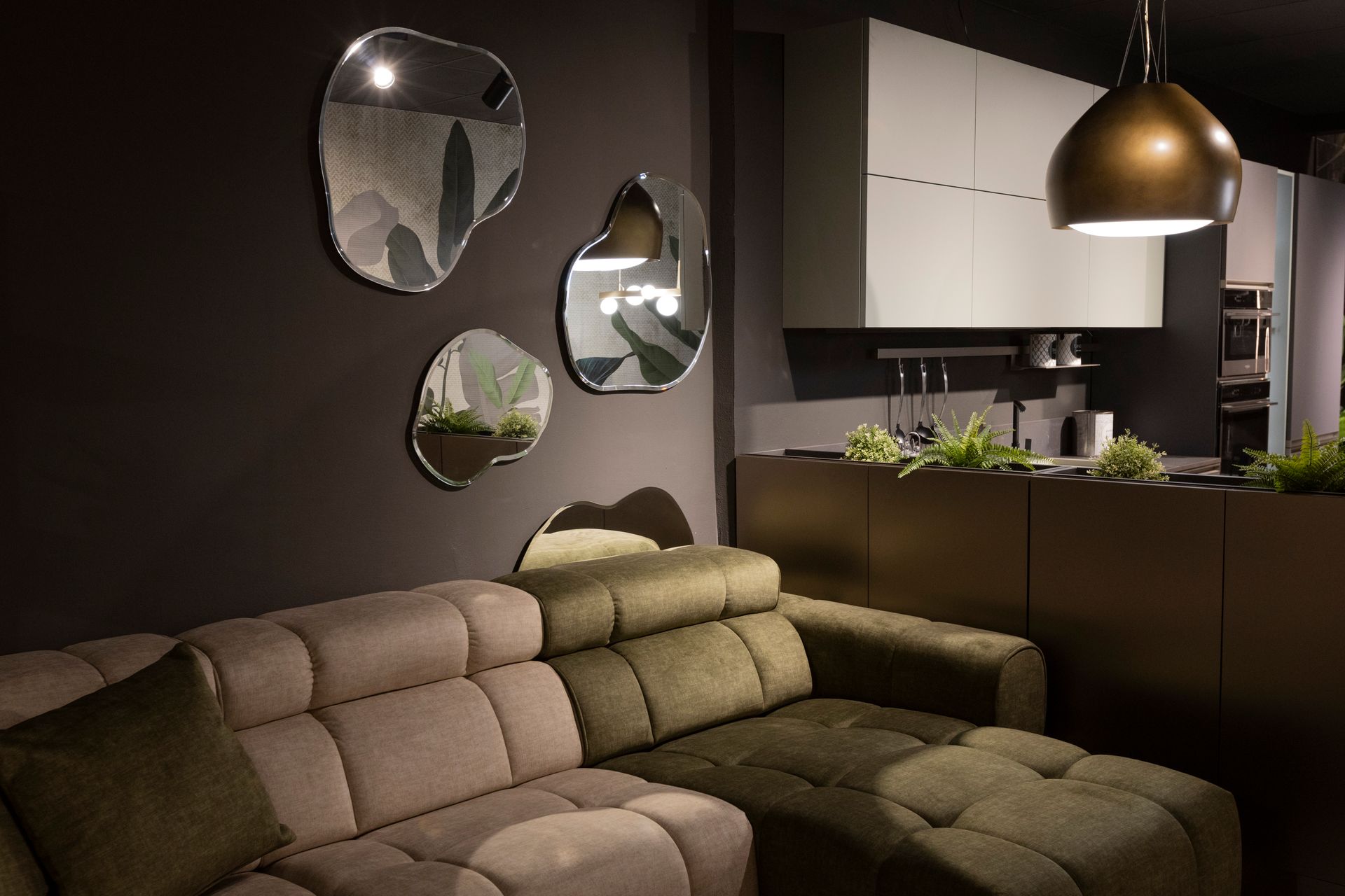 Soggiorno con divano componibile verde oliva, specchi astratti e cucina sullo sfondo.