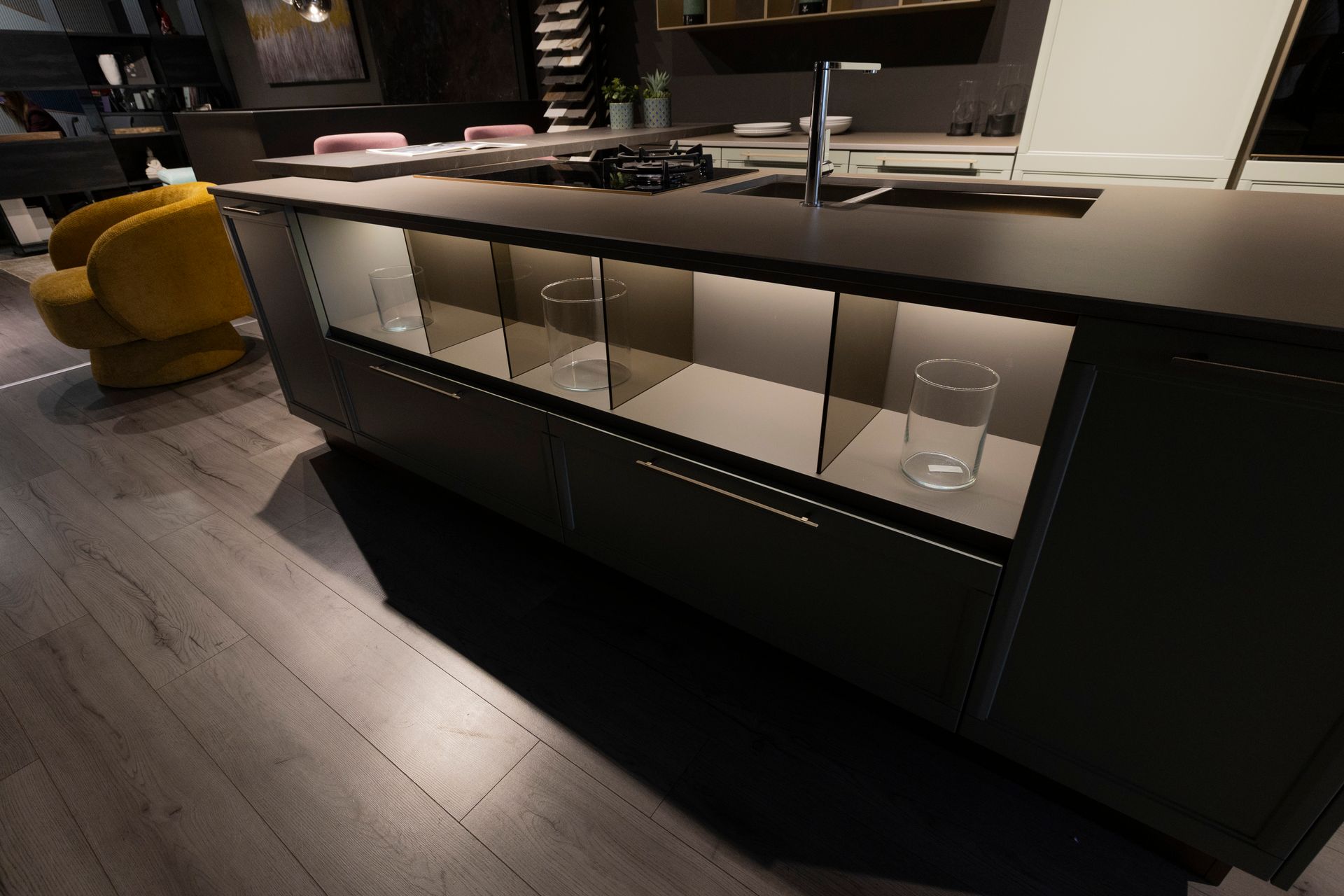 Isola cucina con ripiani in vetro integrati. Mobili scuri, piano di lavoro grigio. Pavimento in legno, poltrona gialla a vista.