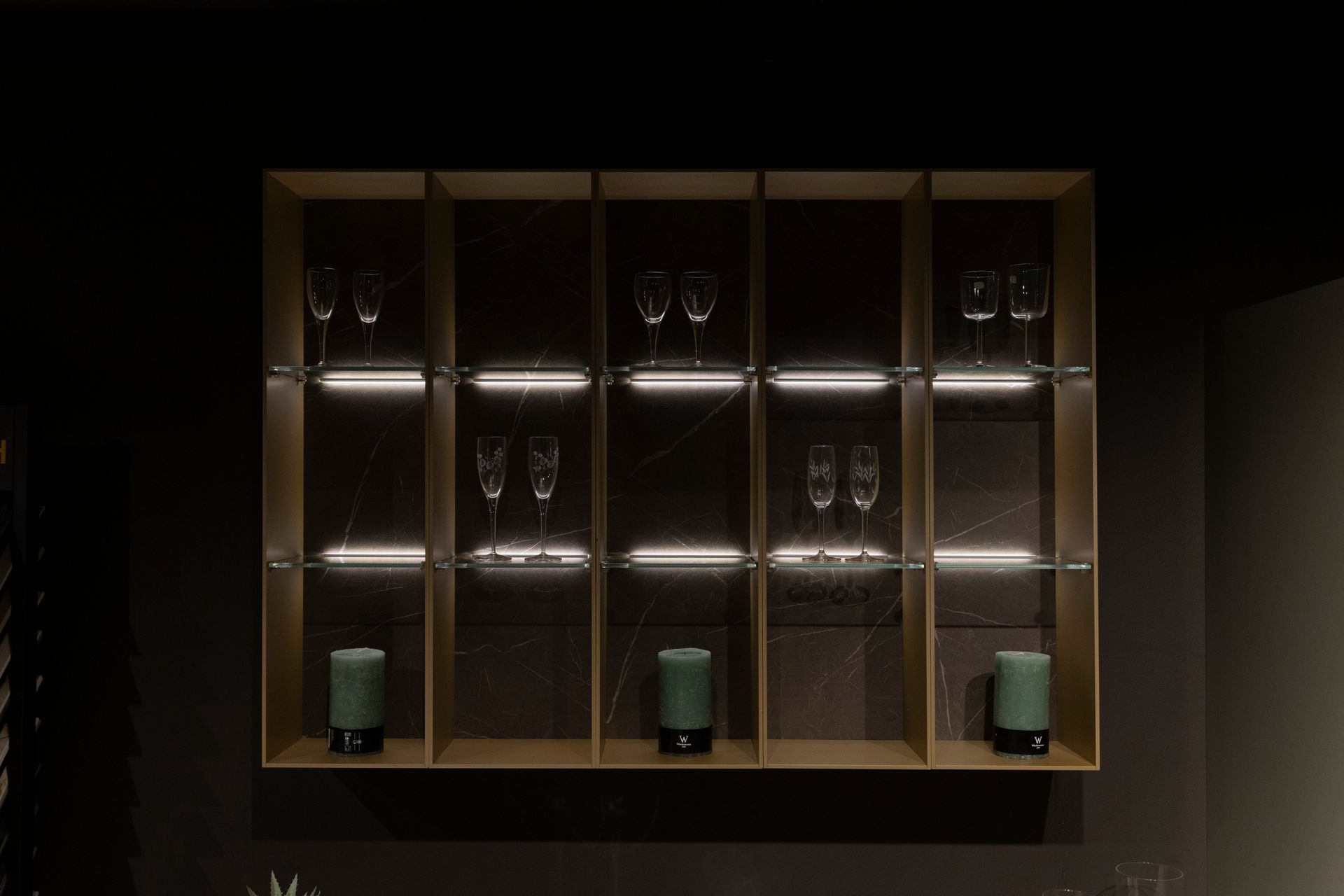 Vetrina illuminata incorporata, con esposizione di bicchieri e candele cilindriche verdi contro una parete scura.