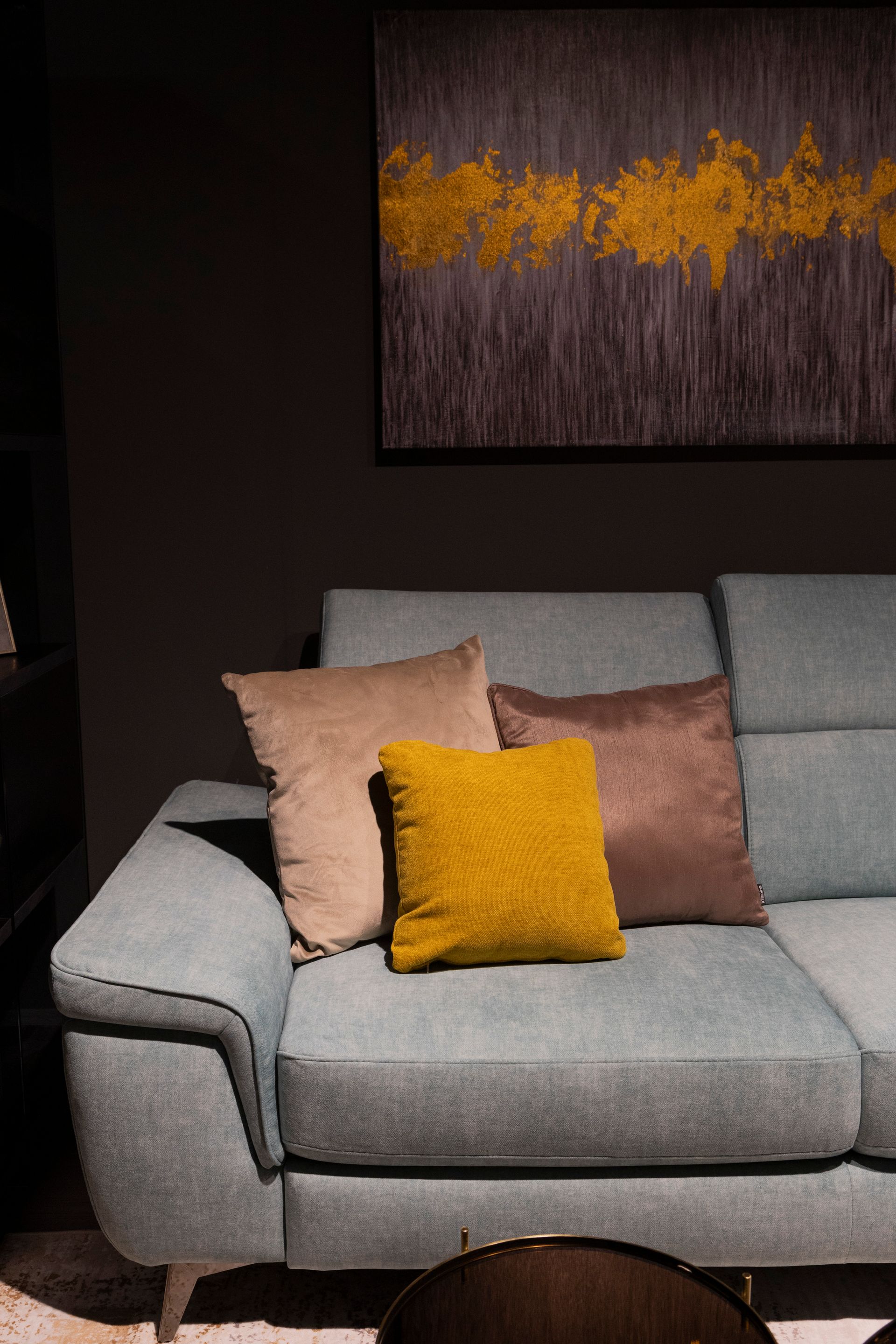 Divano blu con cuscini marroni, gialli e beige, sotto un dipinto astratto color oro e grigio.