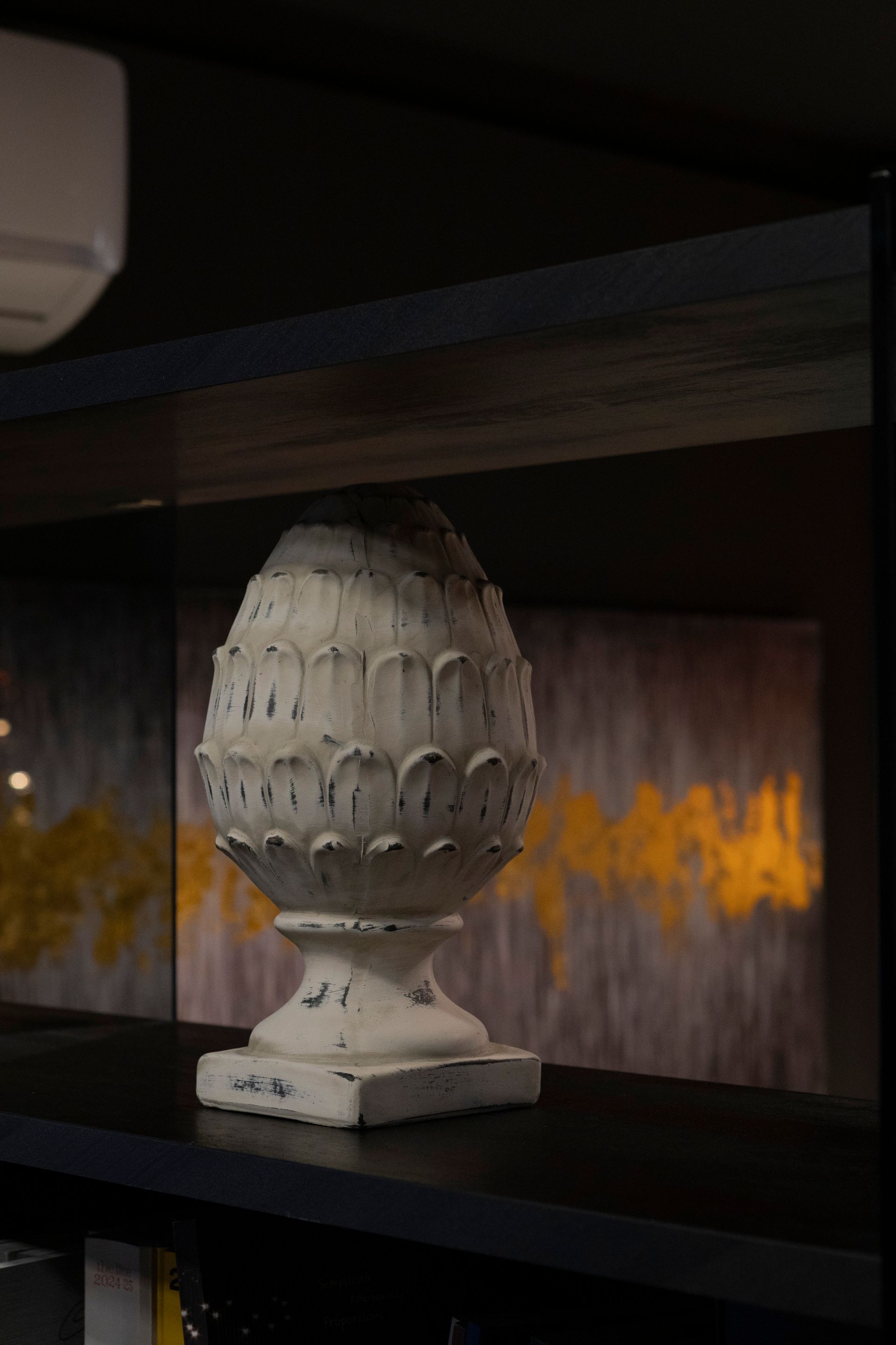 Scultura decorativa a forma di pigna bianca e consumata dal tempo su una mensola, con decorazioni gialle e dorate sullo sfondo.