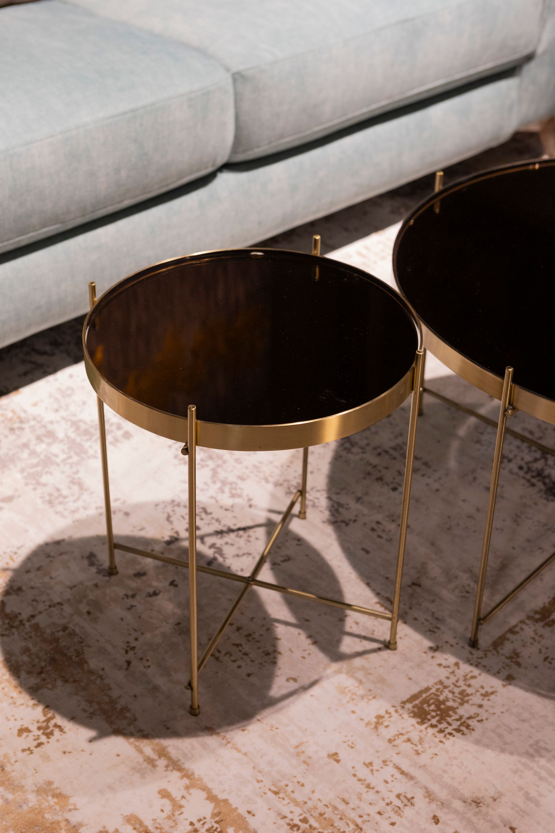 Due tavolini rotondi con cornice dorata e piano marrone scuro, vicino a un divano azzurro su un tappeto a fantasia.