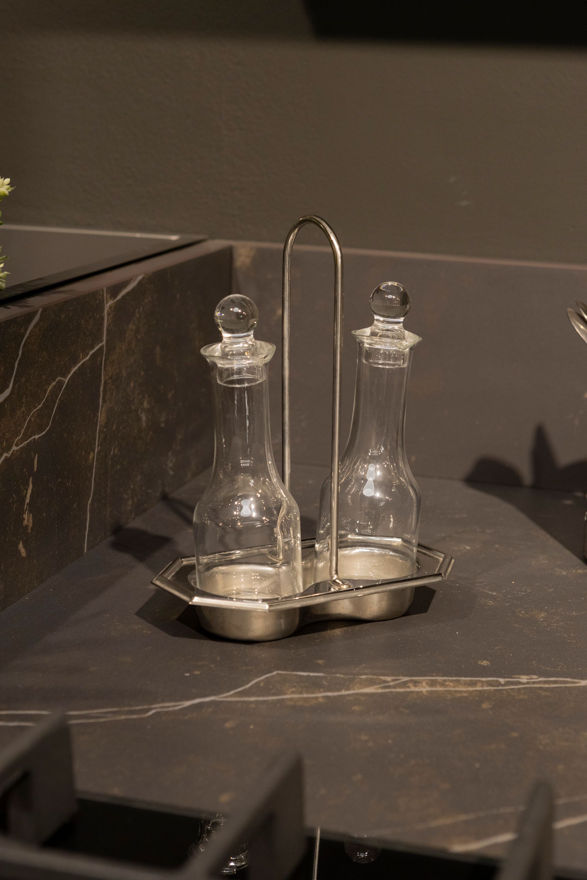 Bottiglie di vetro trasparente per olio e aceto in un contenitore d'argento su un piano di lavoro scuro.