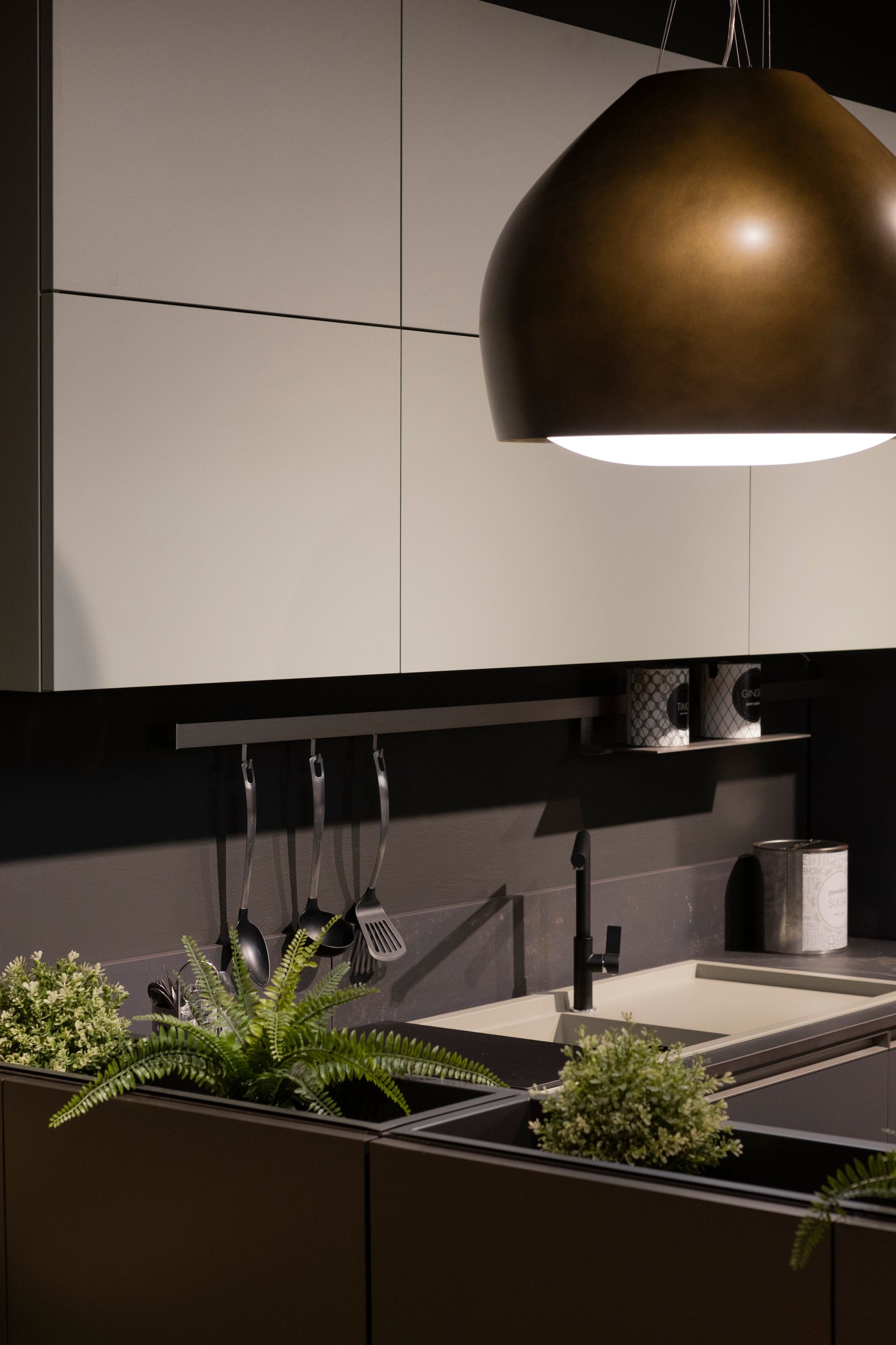 Cucina moderna con lampada in bronzo sospesa, mobili chiari, ripiani e paraschizzi neri e piccole piante.