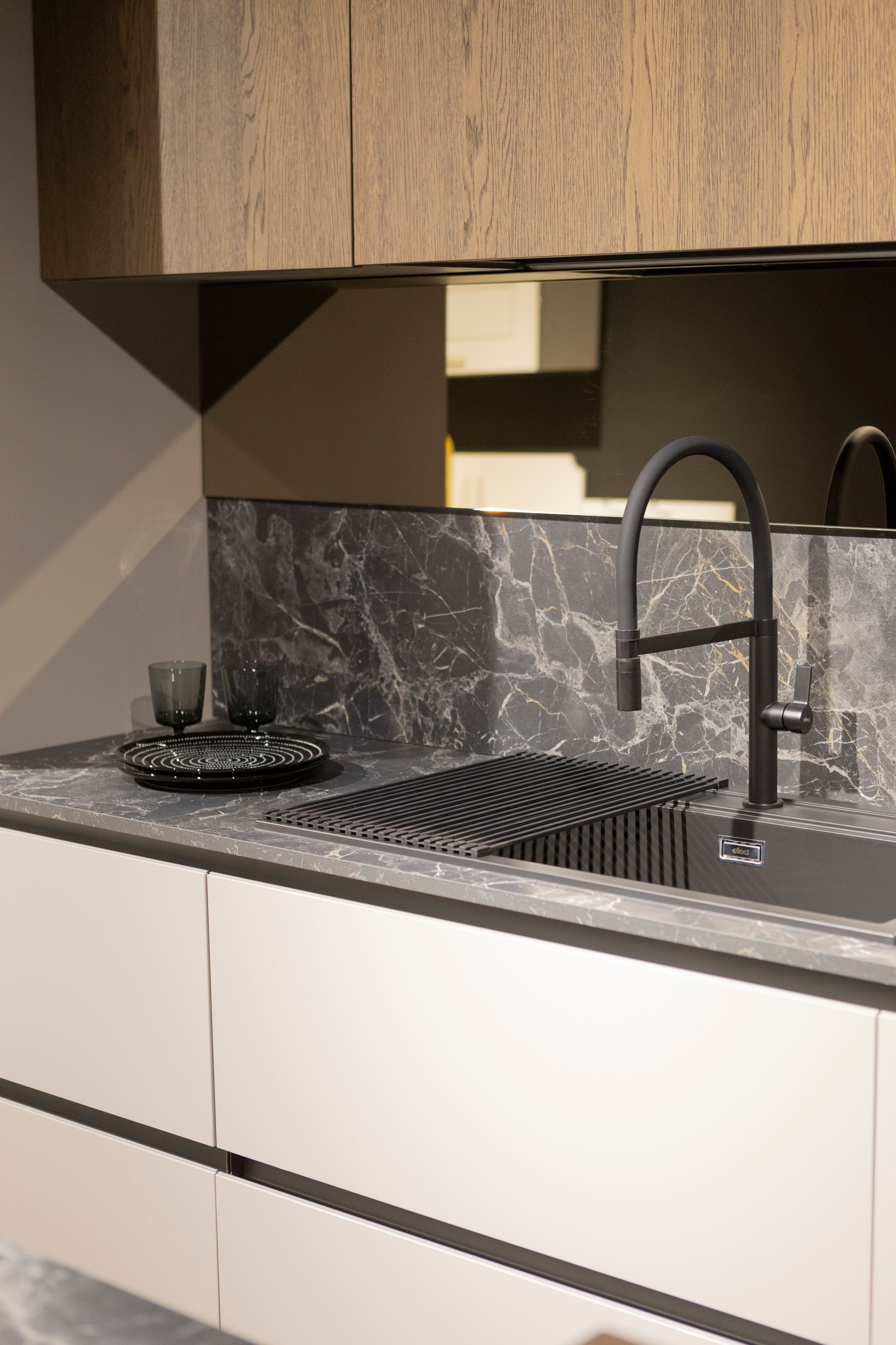 Cucina moderna con paraschizzi in marmo scuro, rubinetto nero opaco e mobili chiari.