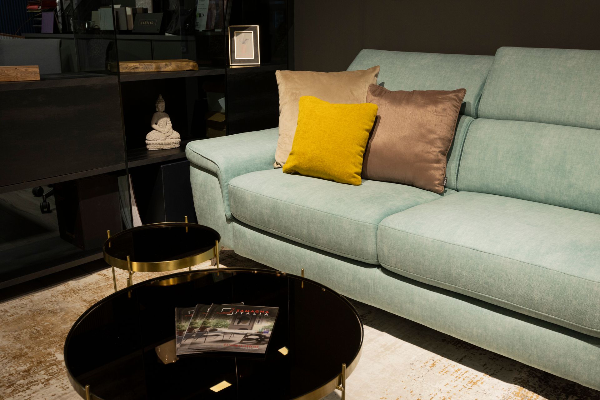 Divano color verde acqua con tre cuscini decorativi, tavolini neri e una libreria scura.
