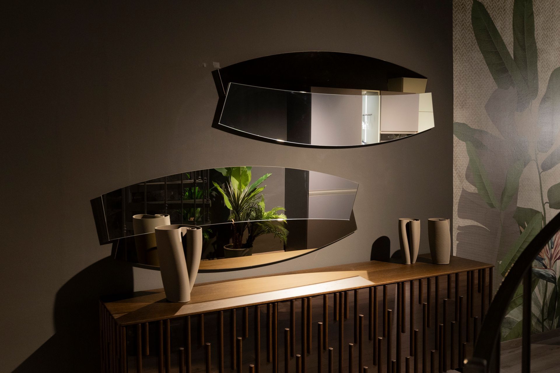 Due specchi moderni sovrapposti sopra un mobile in legno con vasi, contro una parete grigio scuro e carta da parati floreale.