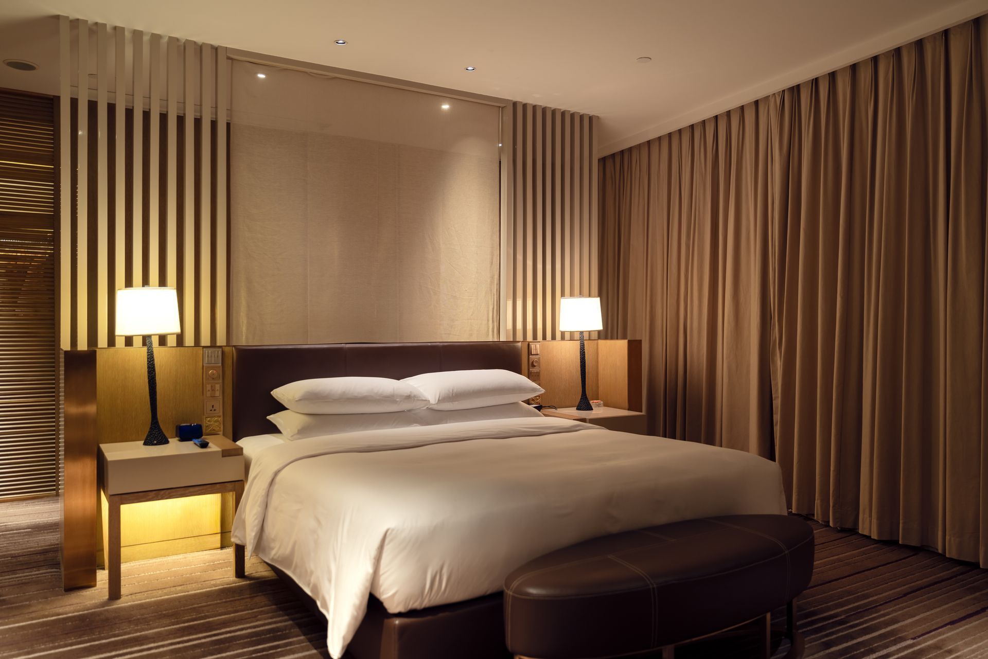 Accogliente camera d'albergo con letto, comodini, lampade e tende. Illuminazione calda, toni neutri.