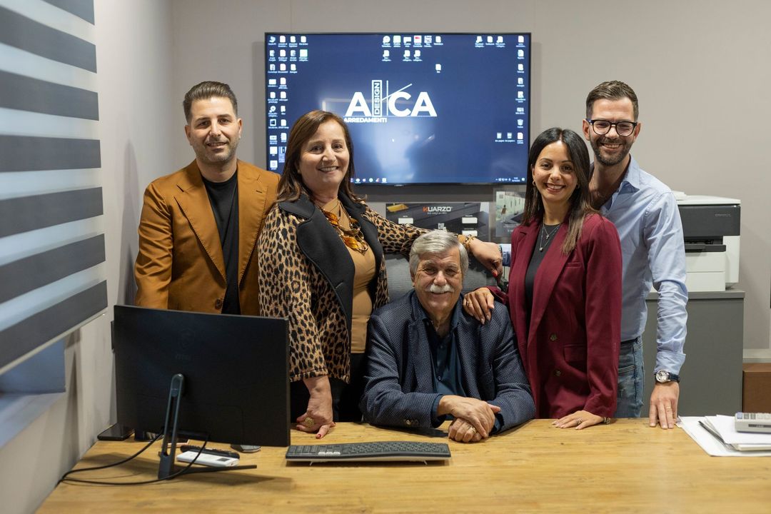 Cinque persone in posa insieme dietro una scrivania in un ufficio con computer e stampante. Sulla parete c'è un logo.