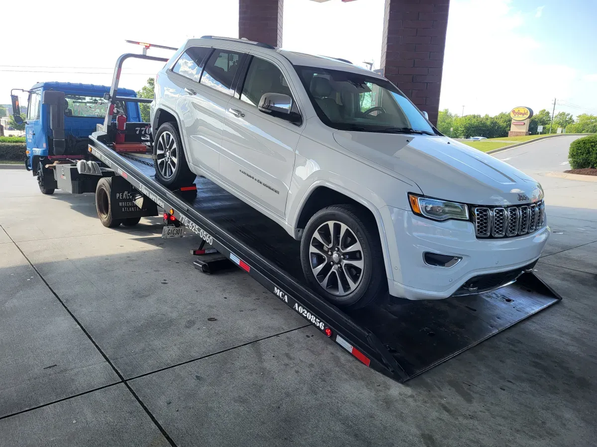 White Jeep SUV being towed on a flatbed truck at a gas station.