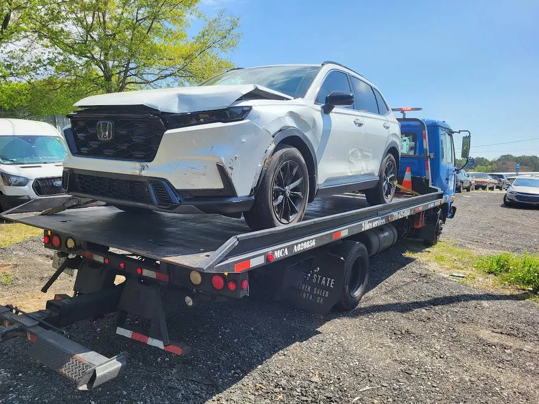 White Honda SUV on a tow truck, damaged front end. Sunny day, gravel lot.