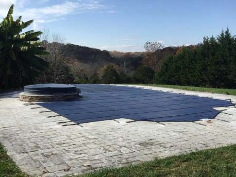Pool covered with a dark blue safety cover, surrounded by stone patio, lawn, and trees under a blue sky.