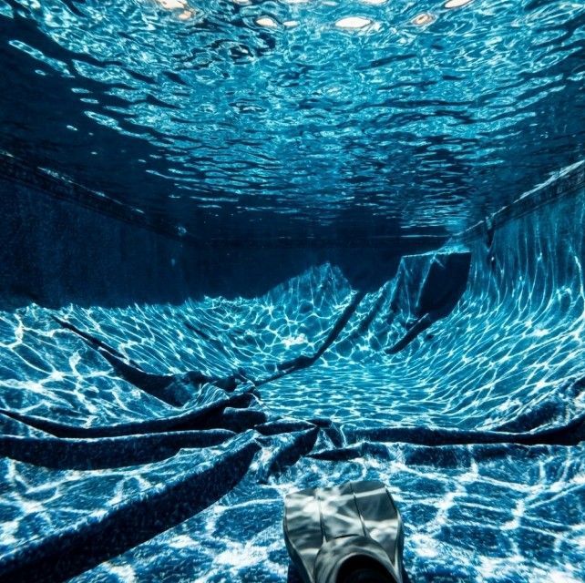 Underwater view of rippling blue water in a pool, with dark figures and a fin visible.