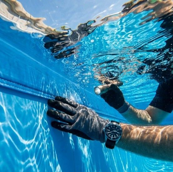 Person underwater, repairing blue pool liner. Black gloves, watch, and flashlight visible.