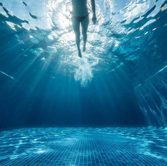 Underwater view of person swimming in a blue-tiled pool, with sunlight streaming down.