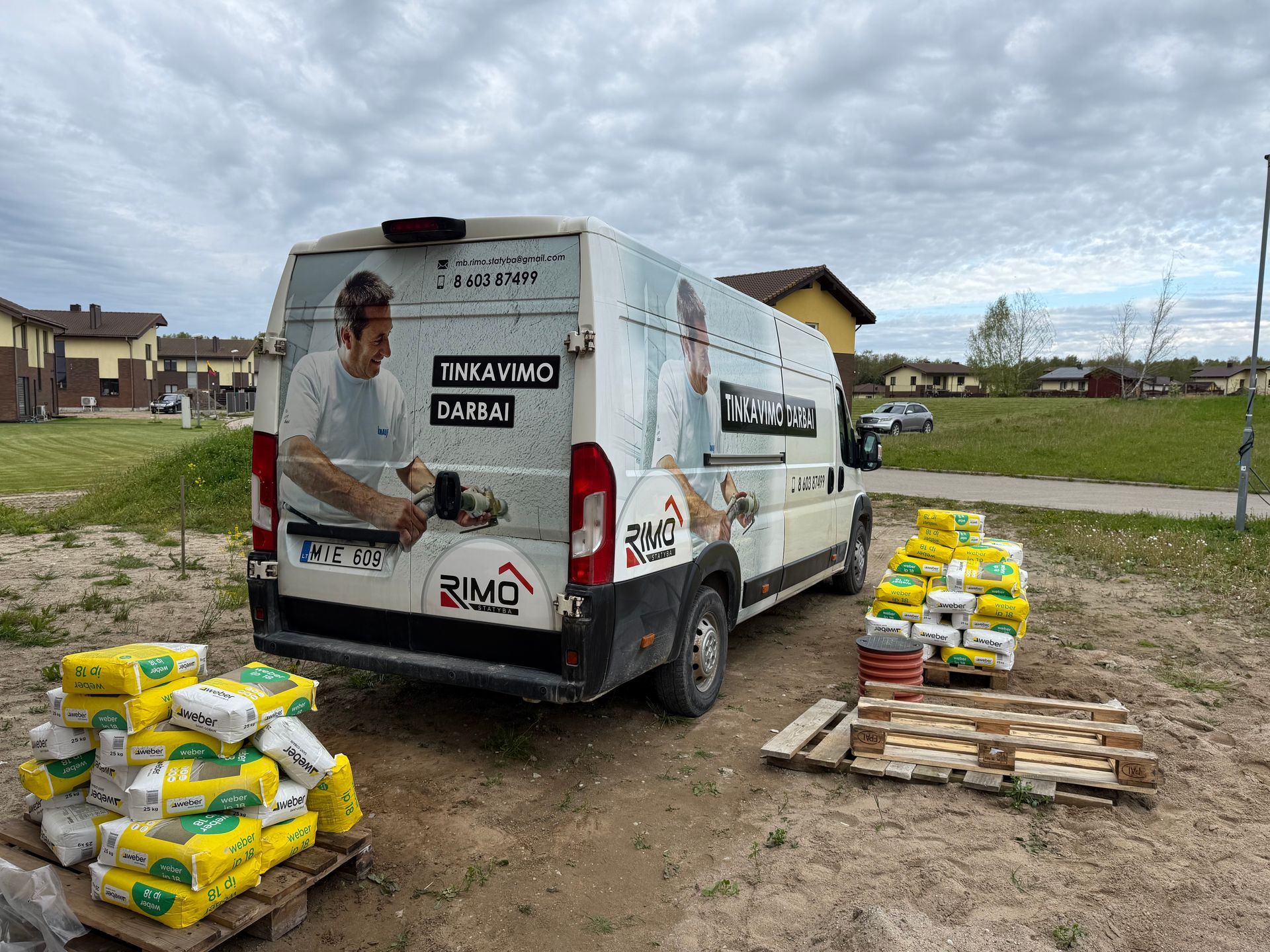 Šalia cemento maišų krūvos stovi baltas furgonas.