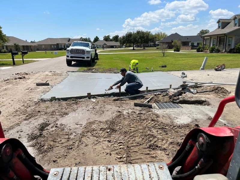 A worker levels wet concrete for a driveway in a residential area; a truck is parked nearby.