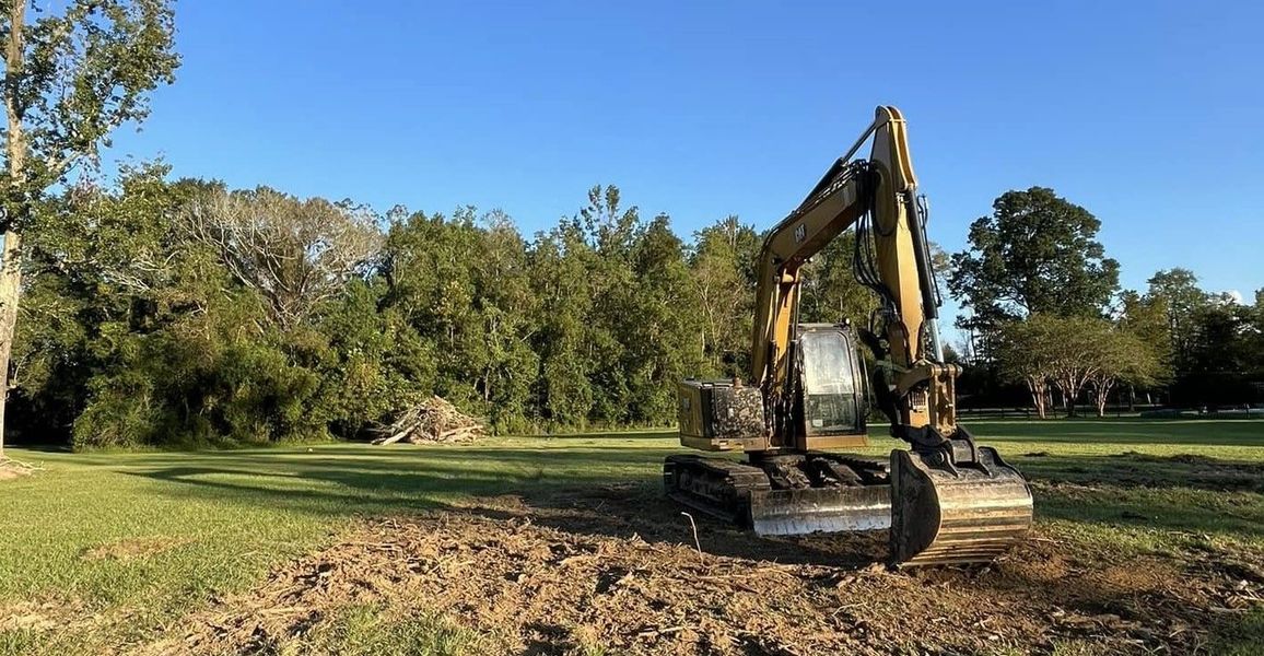 An excavator on a grassy field with trees in the background under a blue sky.