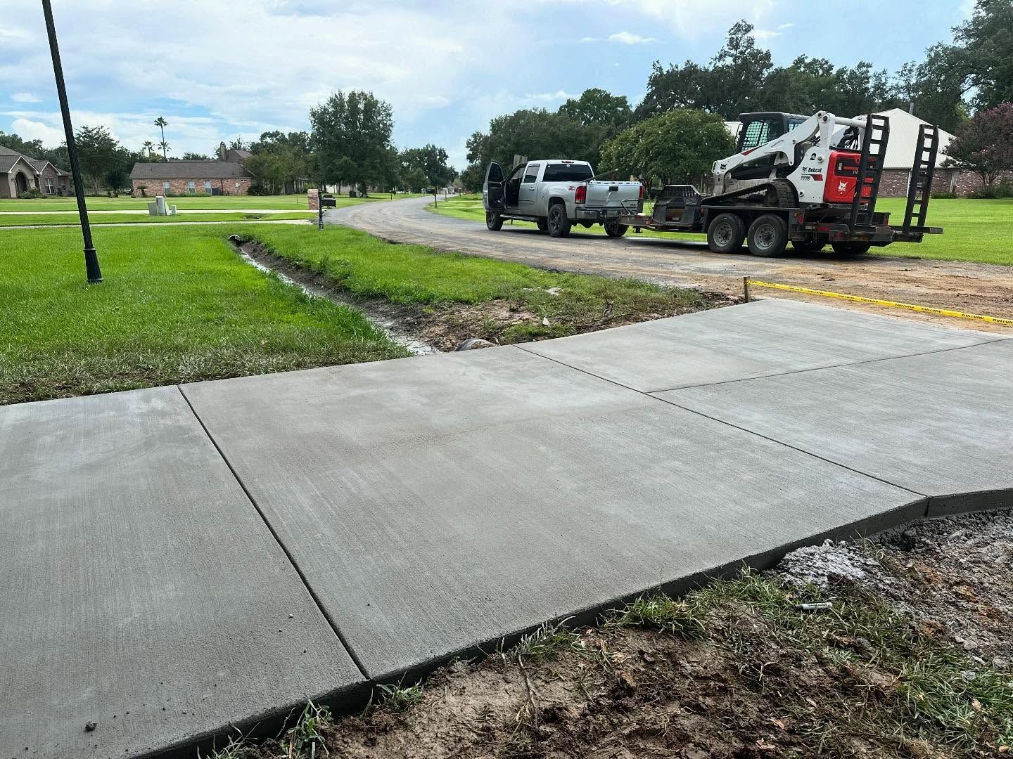 New concrete sidewalk being constructed near a grassy area, with work vehicles in the background.