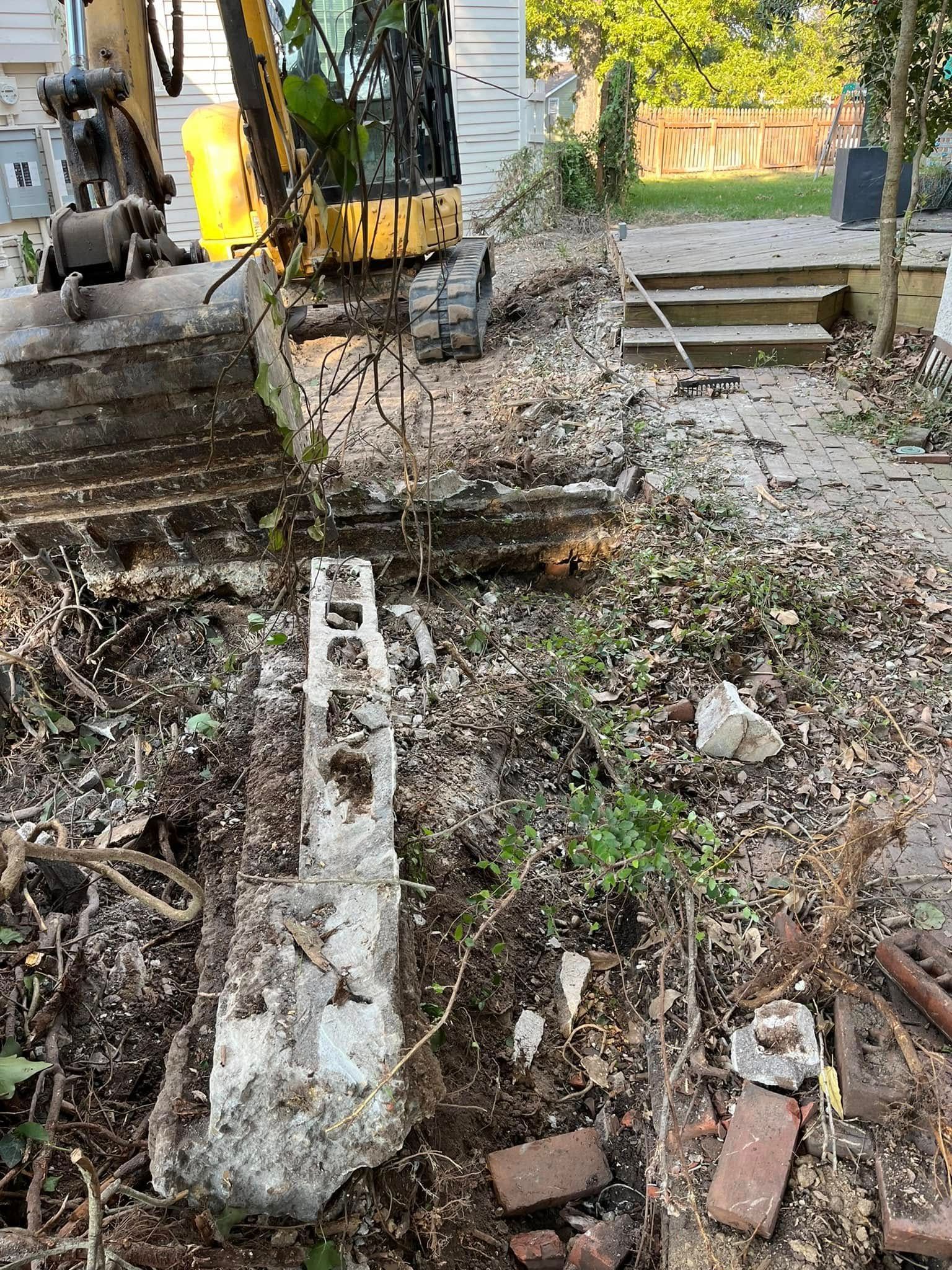 An excavator demolishes a concrete structure in a yard, debris scattered around the work area.