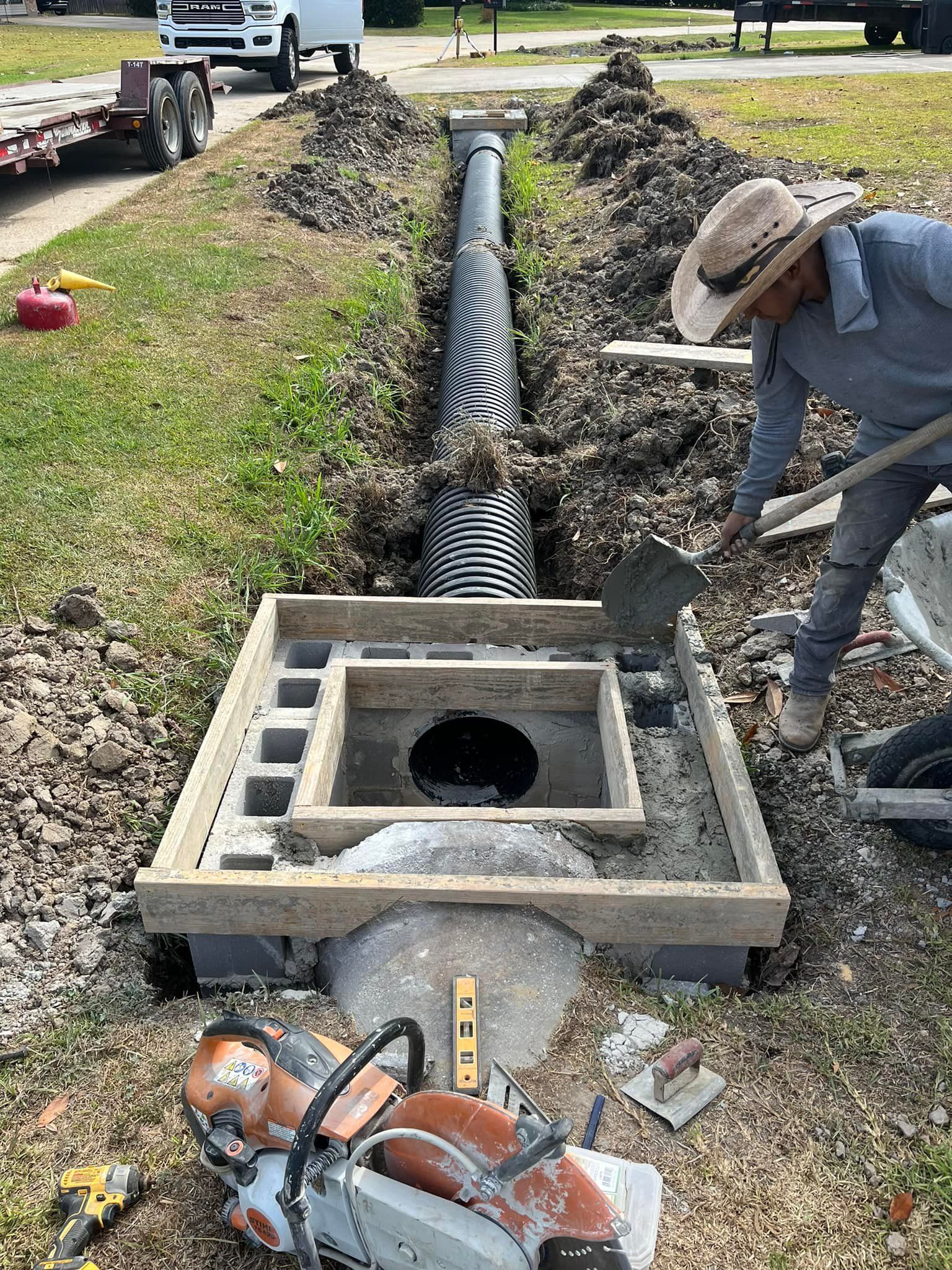 Man shovels concrete into drainage system, with a saw and pipe in a yard.