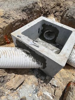 A concrete drainage box with connected corrugated pipes in an open dirt area.