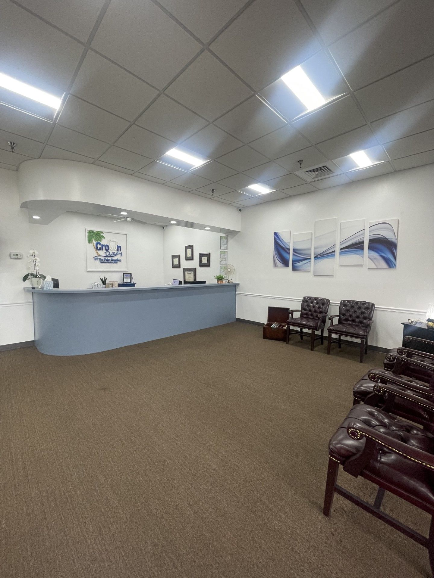 Waiting room with blue reception desk, brown carpet, and chairs. Artwork and overhead lights are present.